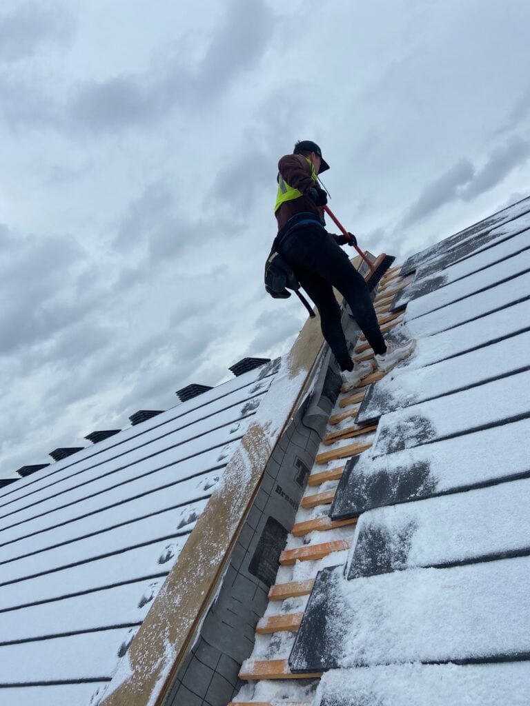 Worker clearing snow from roof with safety gear.