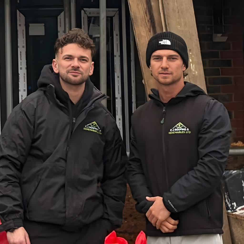 C J Roofing & Renewables co-owners Jack Shaw and Connor Stanley in branded black jackets standing in front of a building.