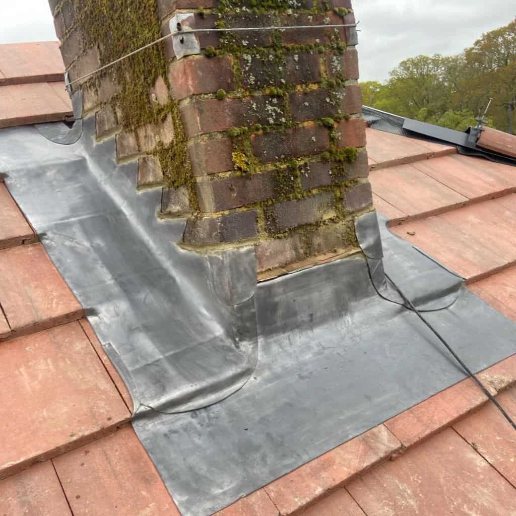 Grey lead flashing dressed around a moss-covered brick chimney stack on a red clay tile roof.