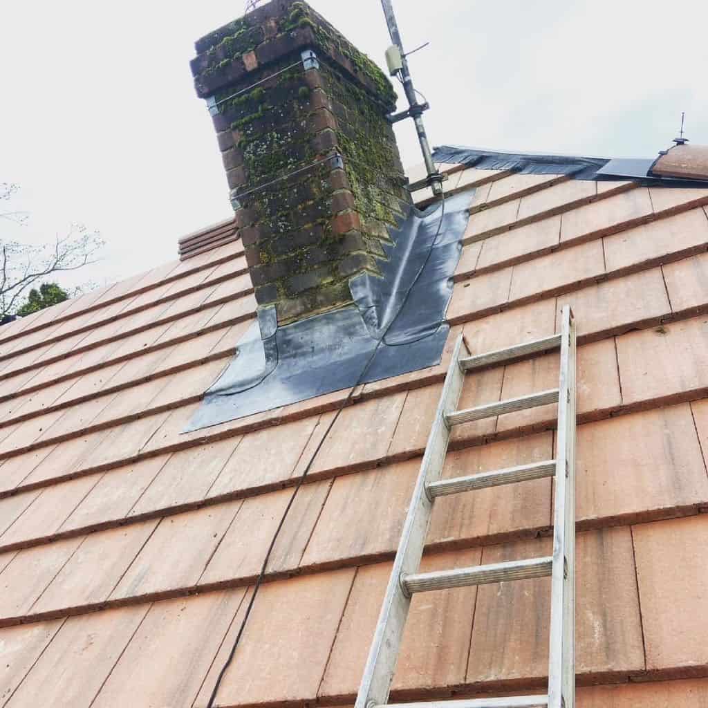 Moss-covered brick chimney with lead flashing on a red clay tile roof, with a ladder leaning against the tiles.