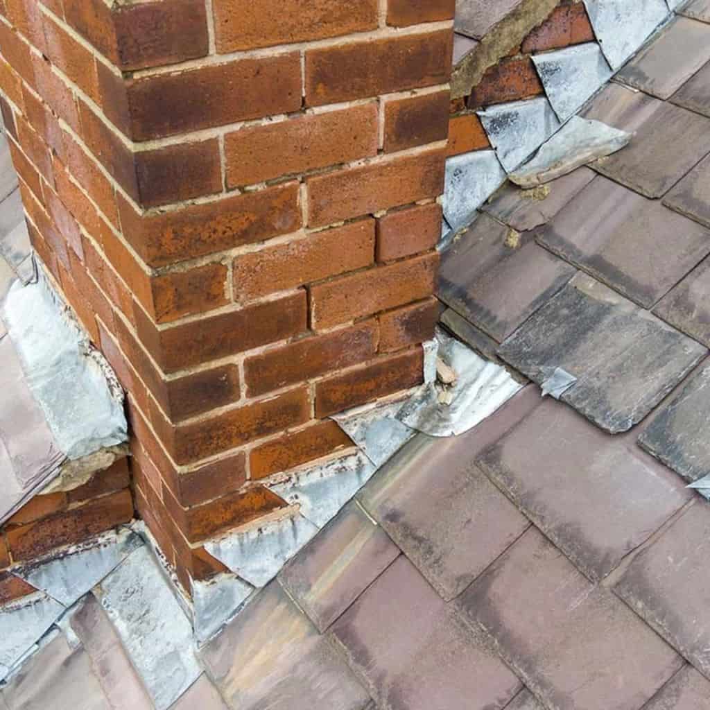 Red brick chimney stack meeting a tiled roof, with weathered lead flashing lifting at the base.