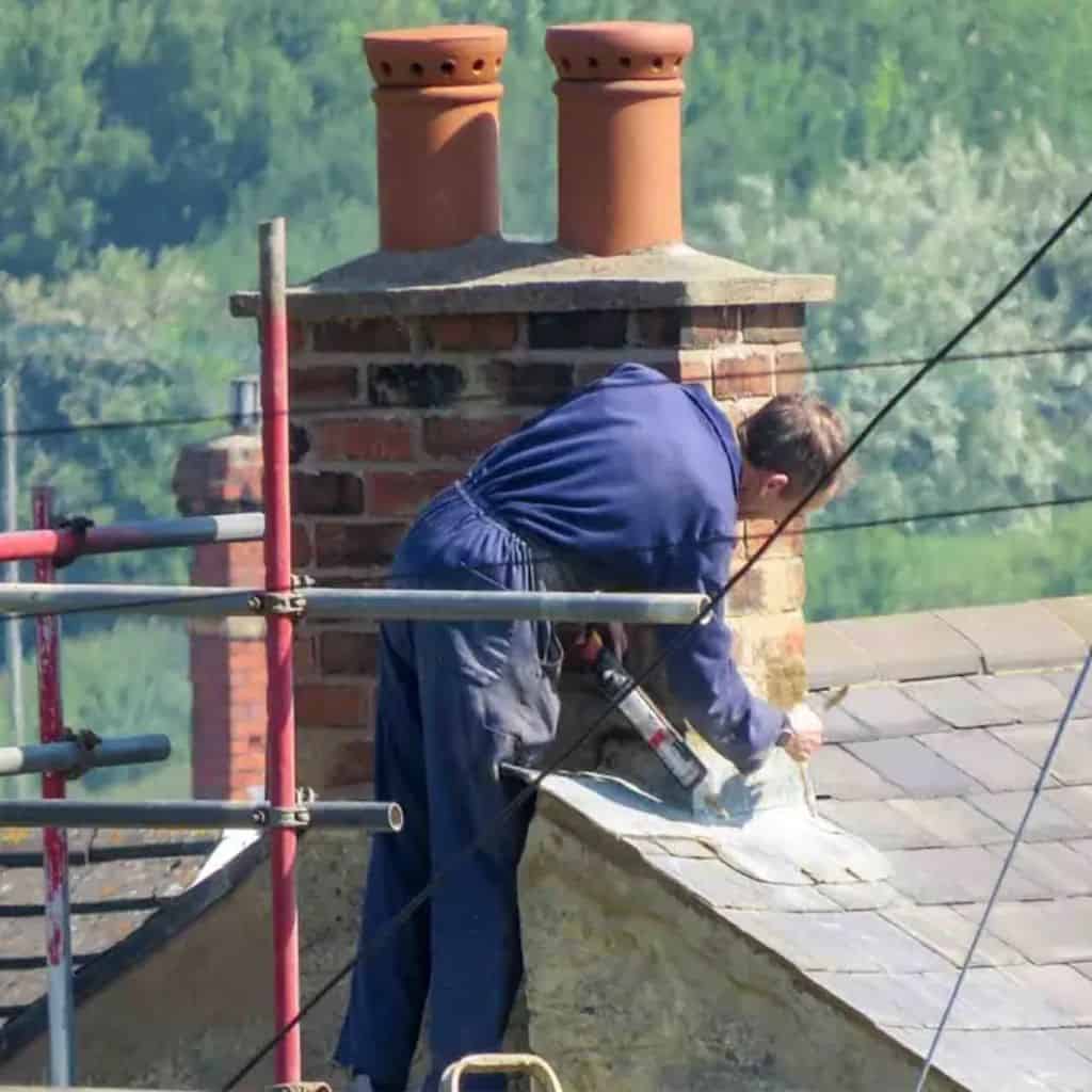 Roofer on scaffolding repairing a brick chimney stack with twin terracotta pots above a slate roof.