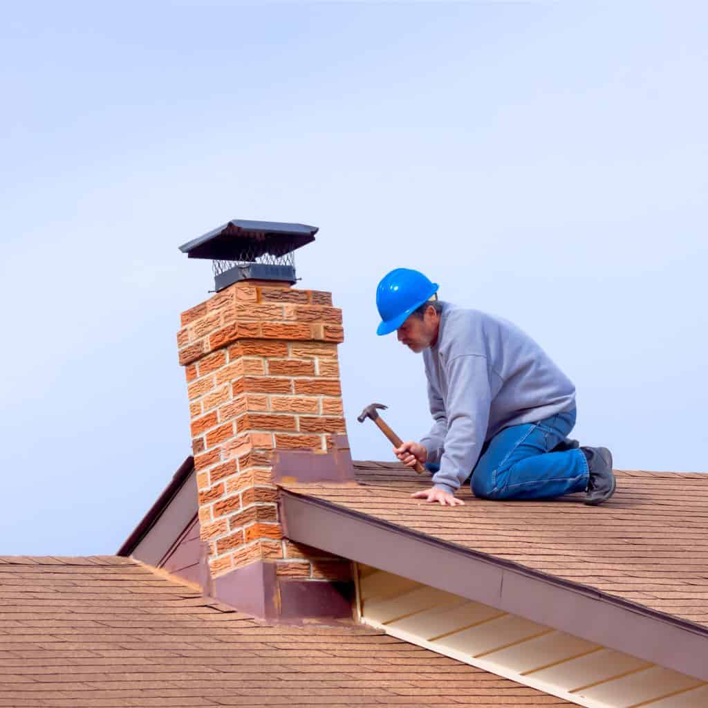Roofer in a blue hard hat kneeling on a shingle roof using a hammer beside a brick chimney stack with a metal cowl.