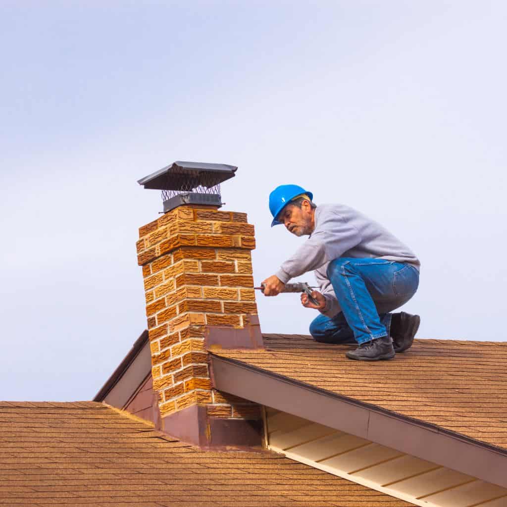 Roofer in a blue hard hat kneeling on a shingle roof working on a brick chimney fitted with a metal cap.