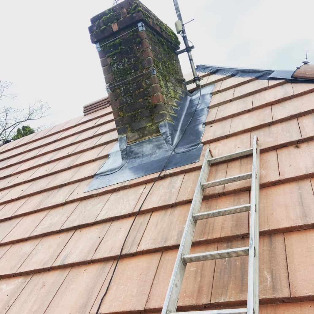 Ladder leaning against a red clay tile roof beside a mossy brick chimney with fresh lead flashing fitted around it.