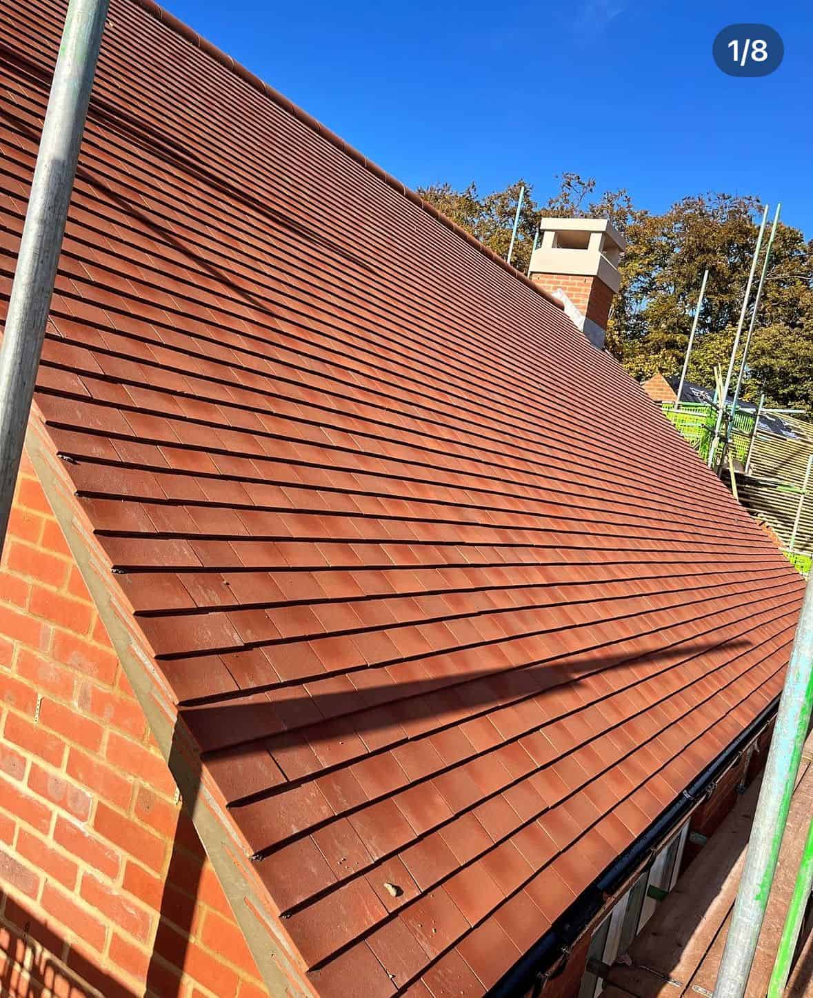 Steep pitched roof of red-brown interlocking tiles meeting a brick gable, with a chimney and trees behind.
