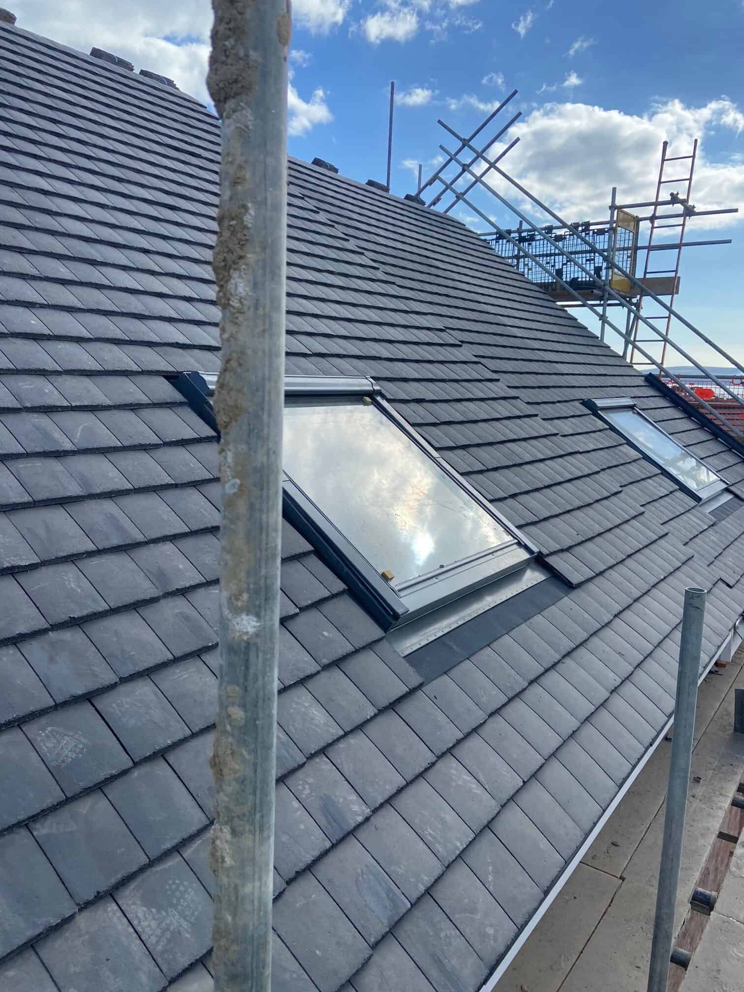 Newly laid grey slate roof with a Velux roof window and scaffolding at the ridge against a bright blue sky.