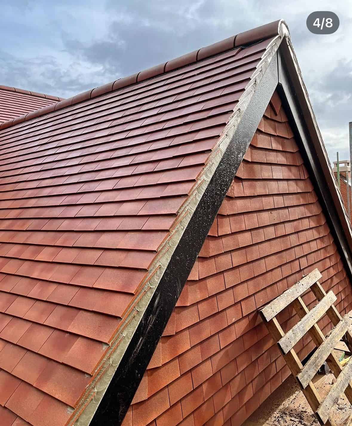 Newly laid red clay plain tile pitched roof with a dry-fix ridge, viewed up the verge against a cloudy sky.