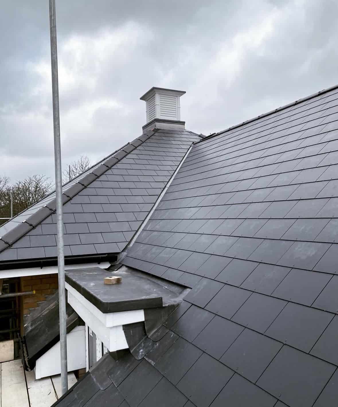Newly laid grey slate-effect tile roof with a hipped ridge, lead valley and white louvred roof vent under a cloudy sky.
