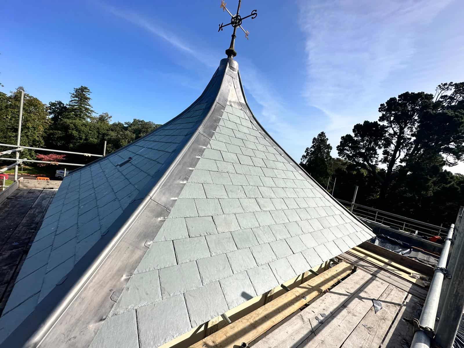 Conical lead and grey slate spire roof topped with an ornate weathervane, surrounded by scaffold boards and trees.