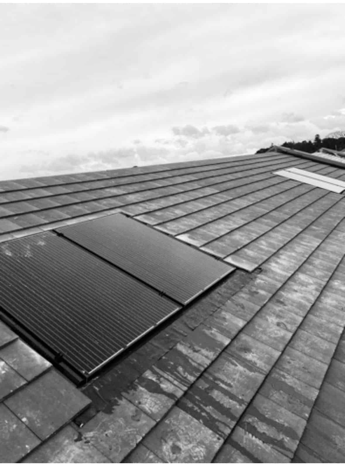 Black-and-white view of black solar panels part-fitted to a tiled roof, with some tiles lifted ready for mounting.