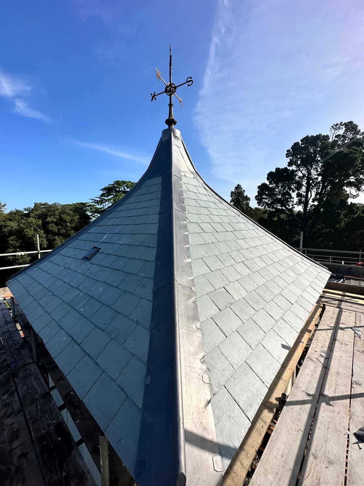 Steeply pitched conical turret roof clad in grey slate with lead detailing and a wrought-iron weathervane against blue sky.