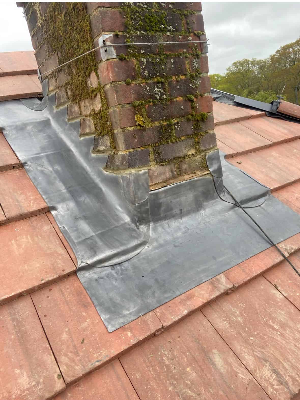 Newly dressed lead flashing around a moss-covered brick chimney set into a red clay tile roof.