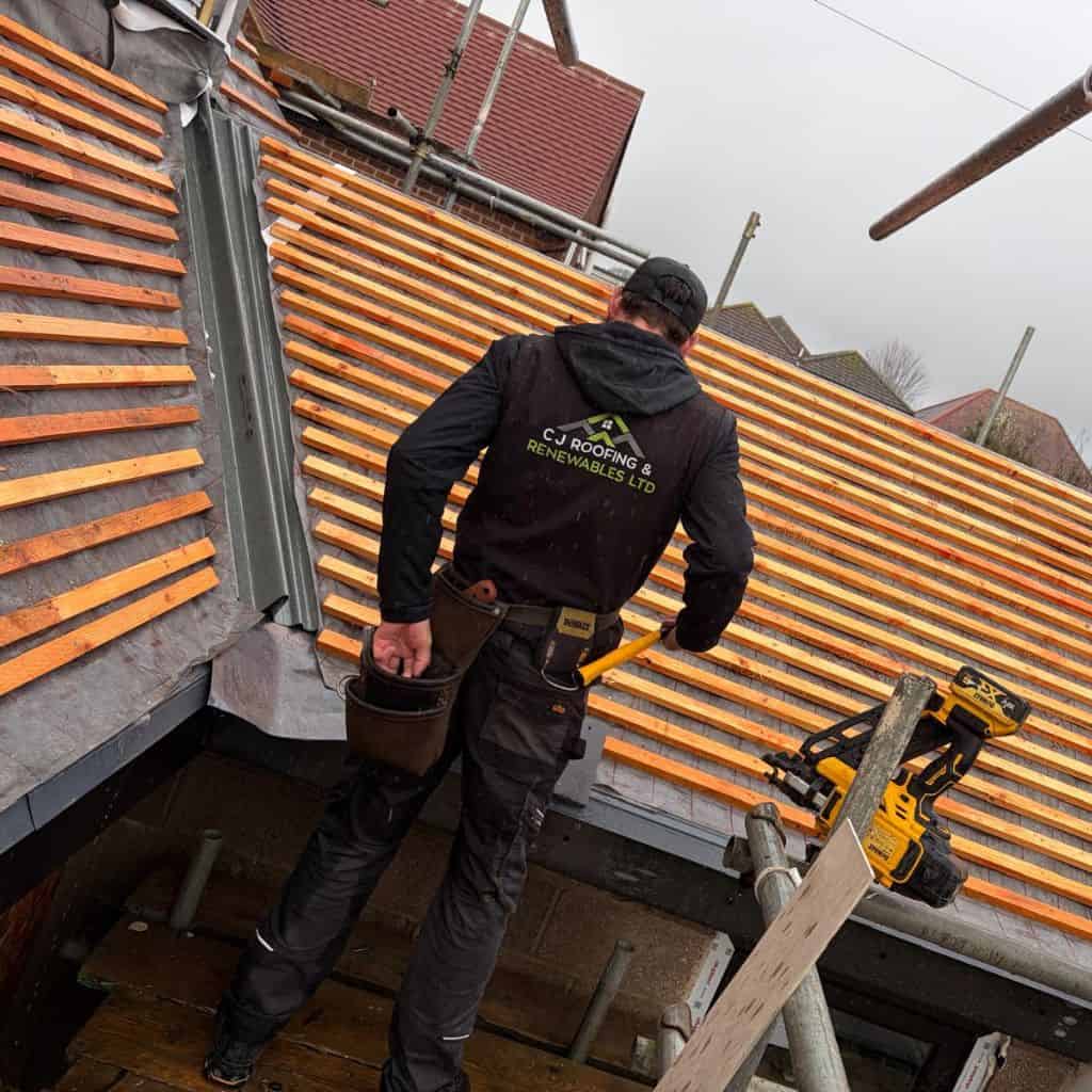 Roofer in branded workwear standing on scaffolding fixing timber battens to a pitched roof being re-tiled.