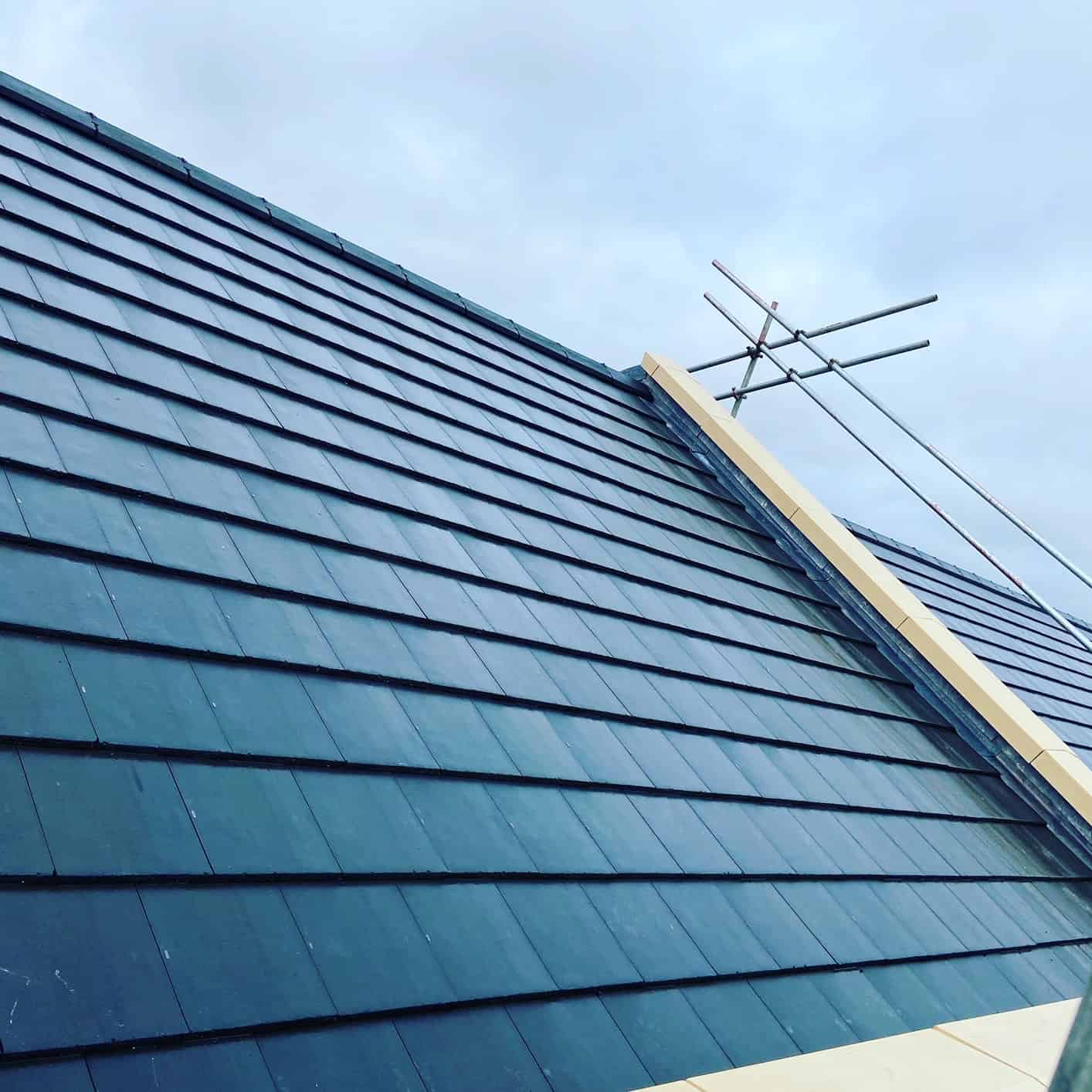 Steeply pitched roof clad in dark grey-blue interlocking slate-effect tiles, with a TV aerial against a cloudy sky.
