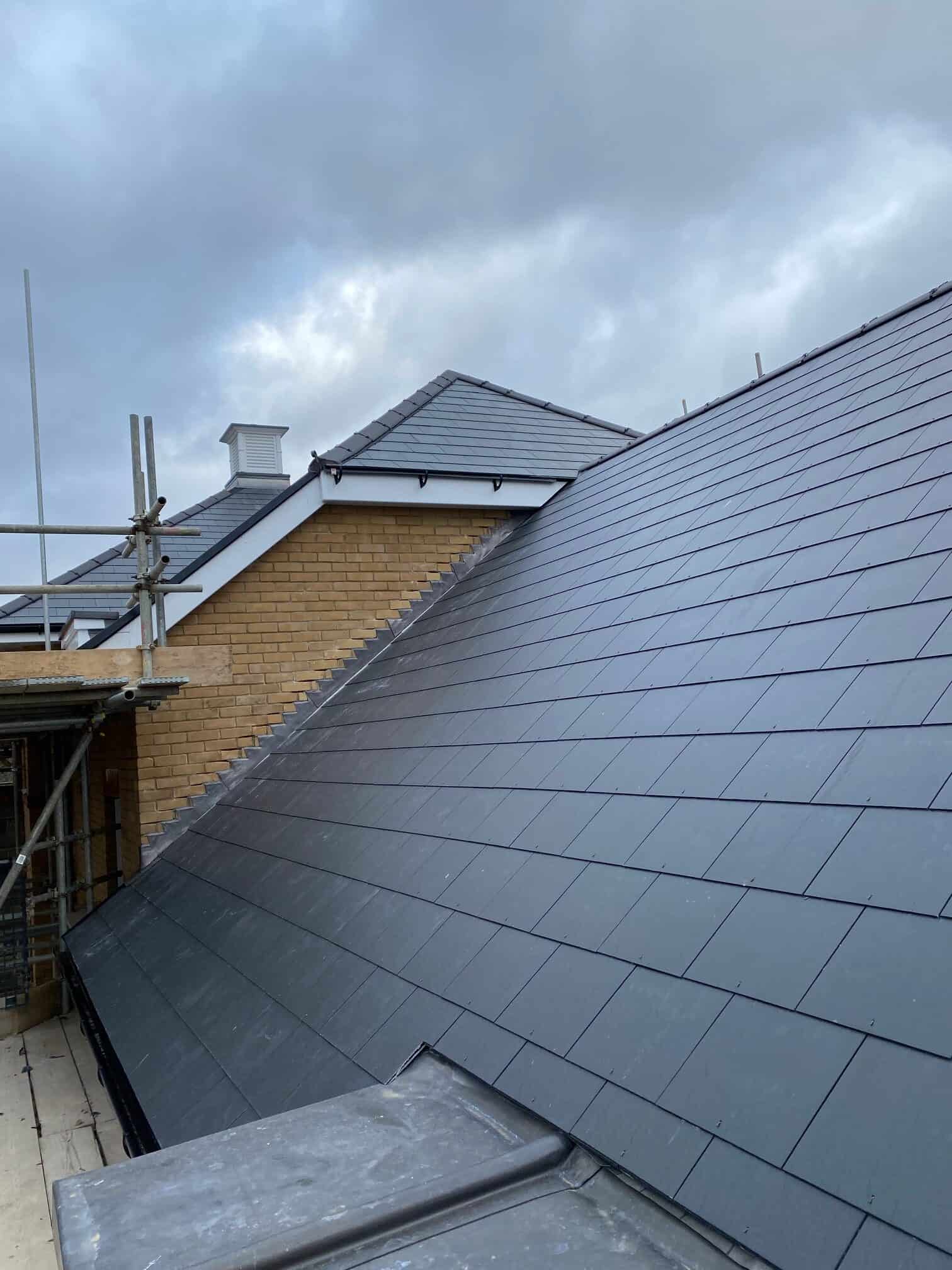 Steep dark grey slate roof on a building with yellow brickwork, scaffolding and a roof lantern under a grey sky.