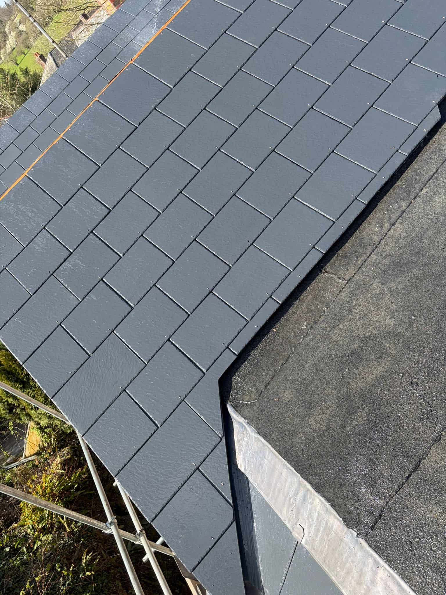 Close-up of newly laid dark grey slate roof tiles meeting a felt-covered flat roof, viewed from scaffolding.