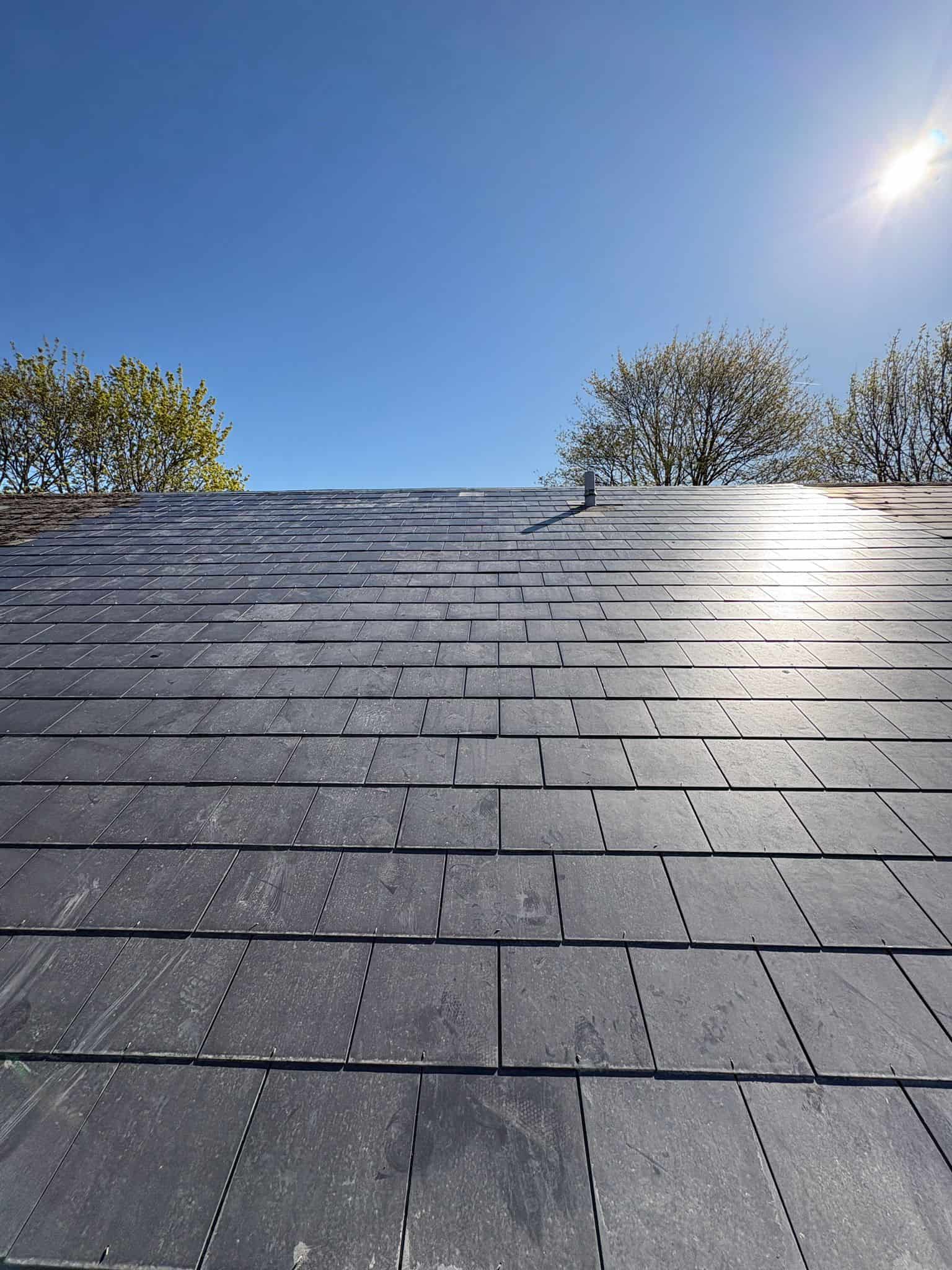 Expanse of dark grey slate tiles on a pitched roof rising to the ridge against a bright blue sky with bare trees.