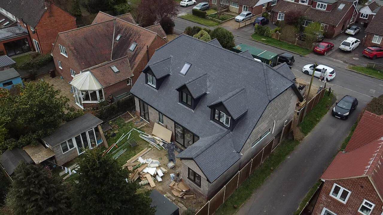 Aerial view of a detached house with a new dark grey tiled roof and three dormer windows, materials in the garden below.