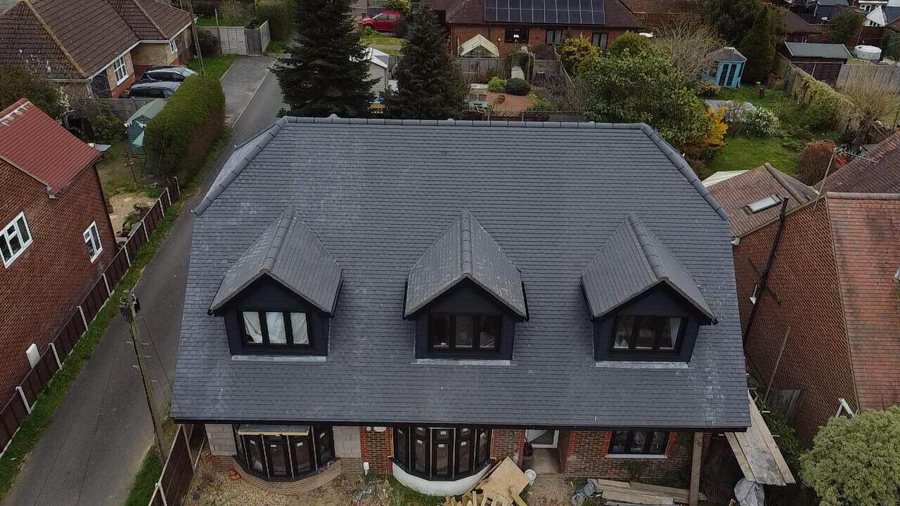 Black solar panels mounted on a grey slate roof beside a red brick chimney stack under a moody, cloudy sky.
