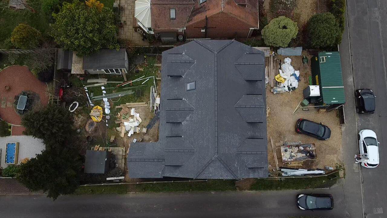 Aerial view of a long grey slate roof on a detached house, with cars and a building site in the surrounding garden.