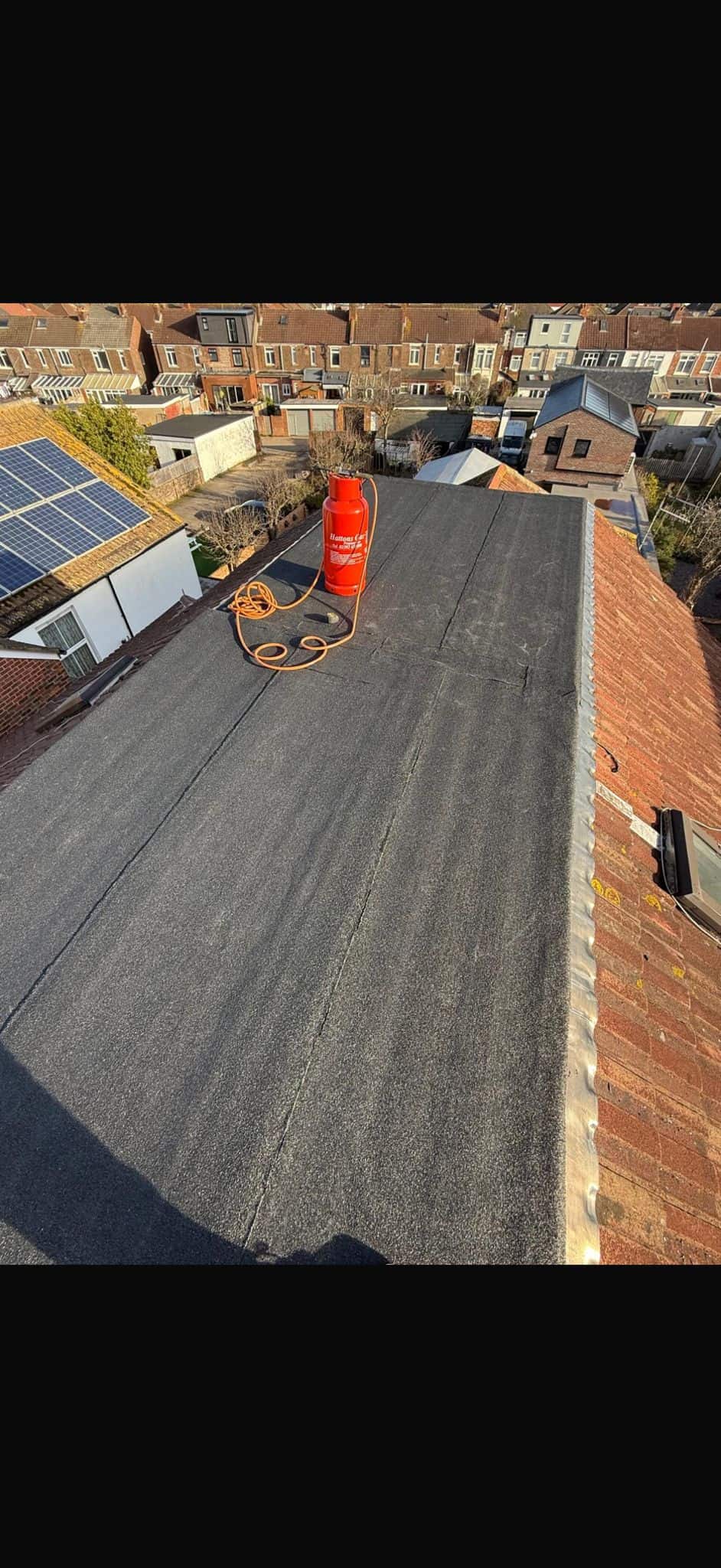 Worker in a red cap kneeling to lay black felt over timber decking on a flat roof beside a building.