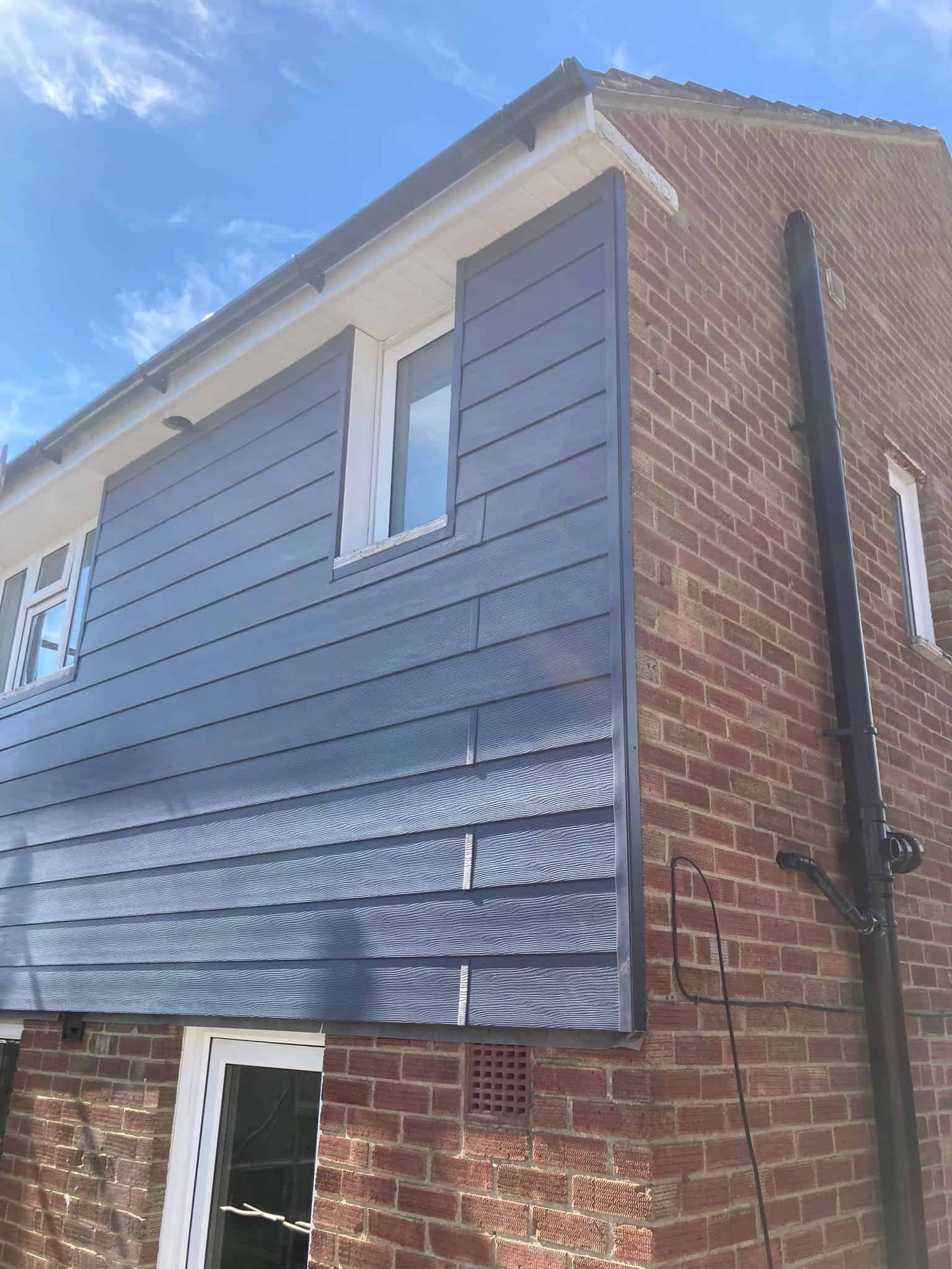 Grey fibre cement weatherboard cladding on the upper storey of a red brick house against a blue sky.