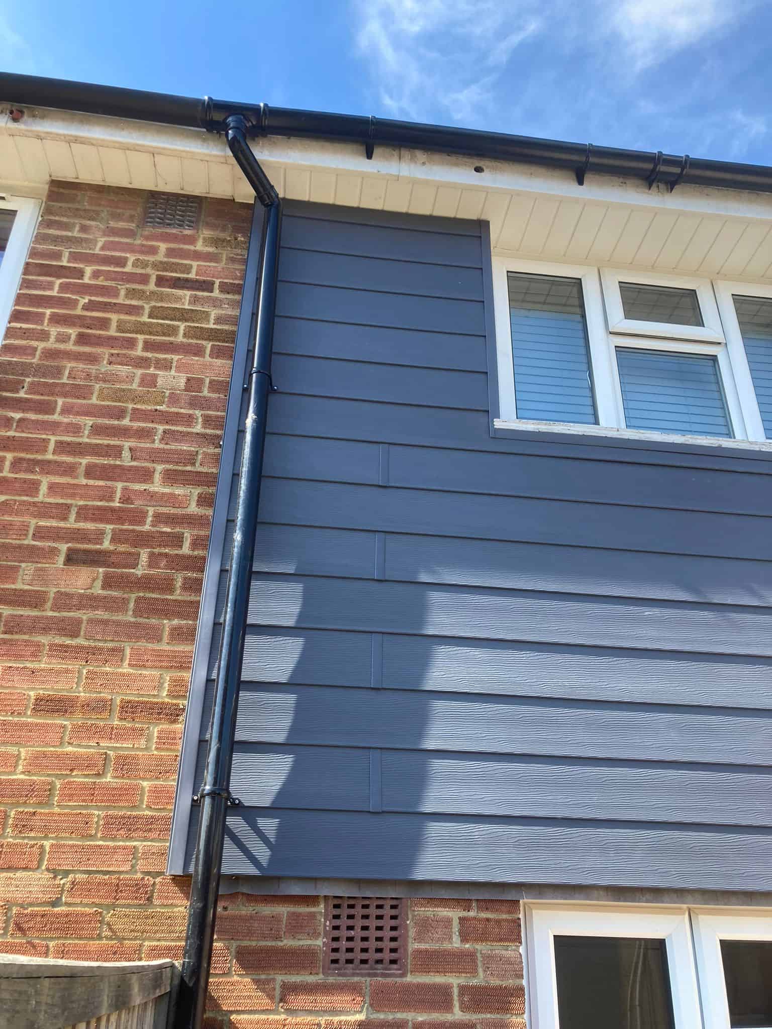 Dark grey horizontal cladding panels fitted to an upper wall beside red brickwork and a black downpipe.