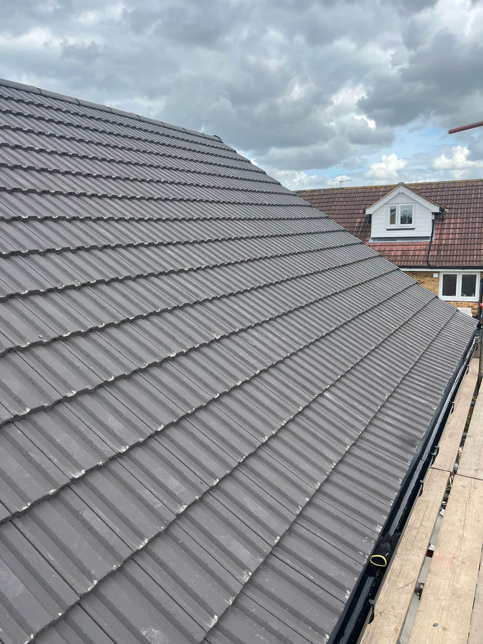 Close view of a freshly laid dark grey tiled pitched roof with neat interlocking rows, a dormer and chimney beyond.