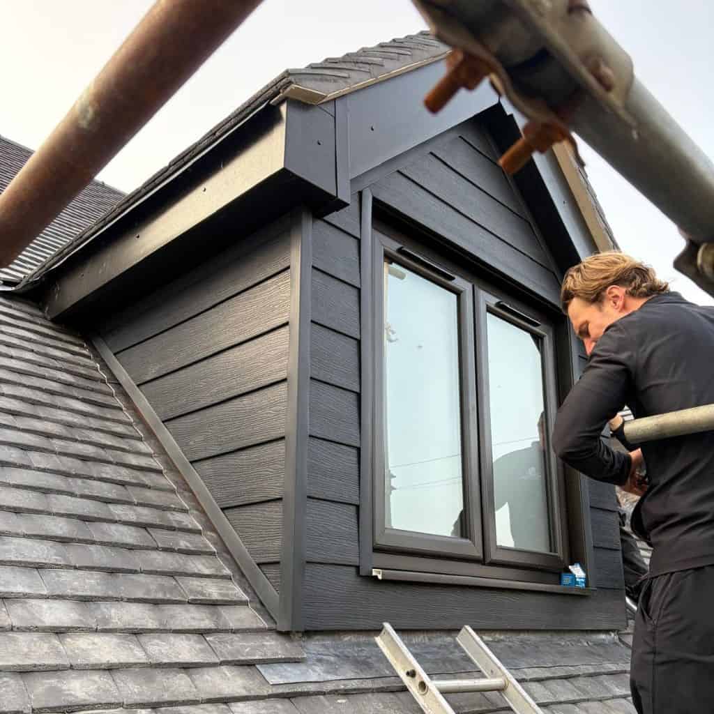 Roofer on scaffolding installing dark fibre cement cladding around a dormer window above a slate roof.