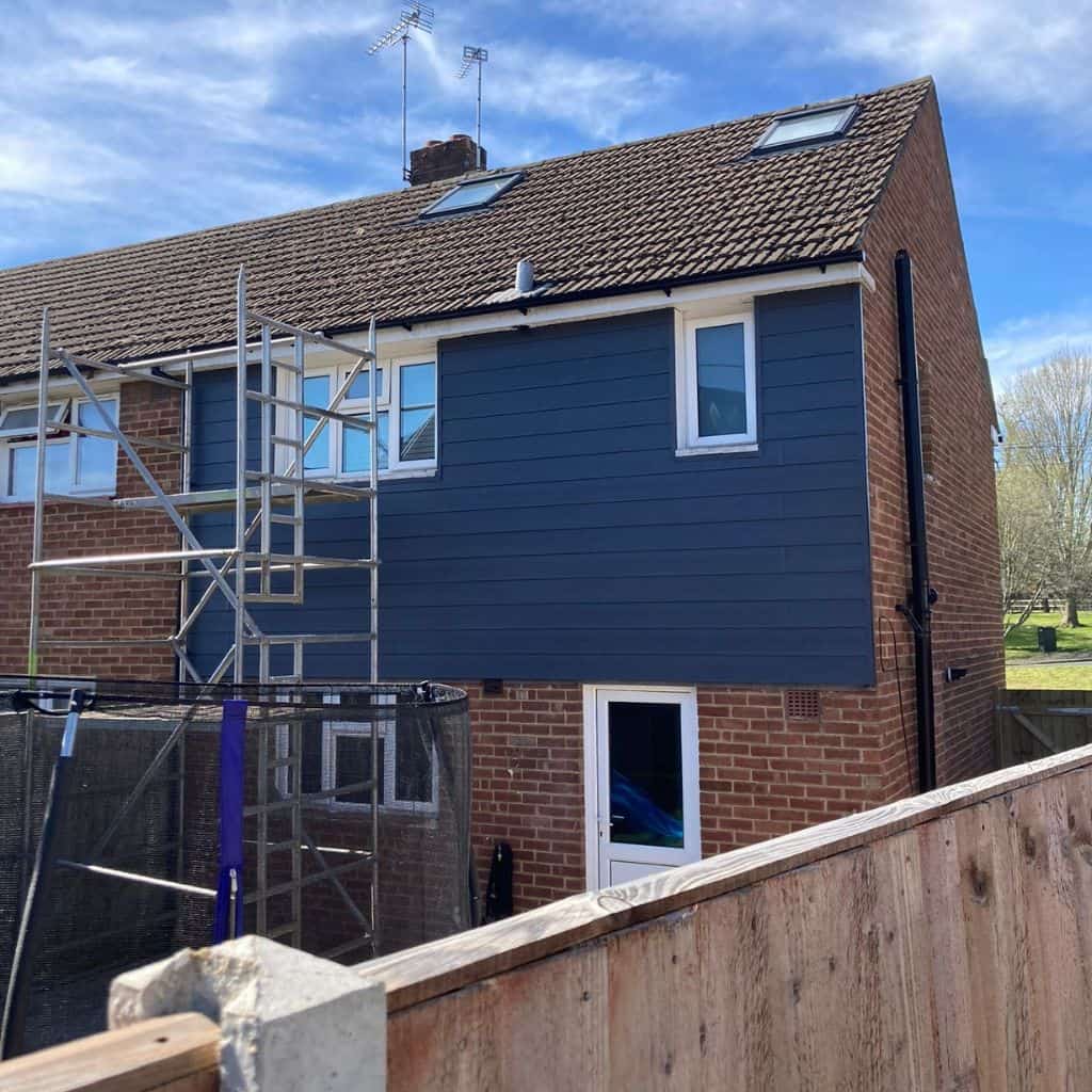 Two-storey brick house with a dark blue weatherboard cladding panel fitted to the upper gable, scaffolding tower to the side.