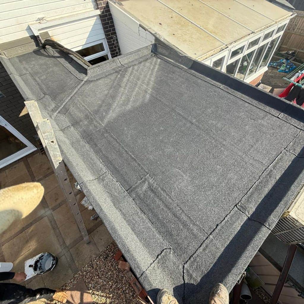 Newly laid grey felt flat roof on a domestic extension, viewed from above beside a conservatory, with a ladder resting on it.