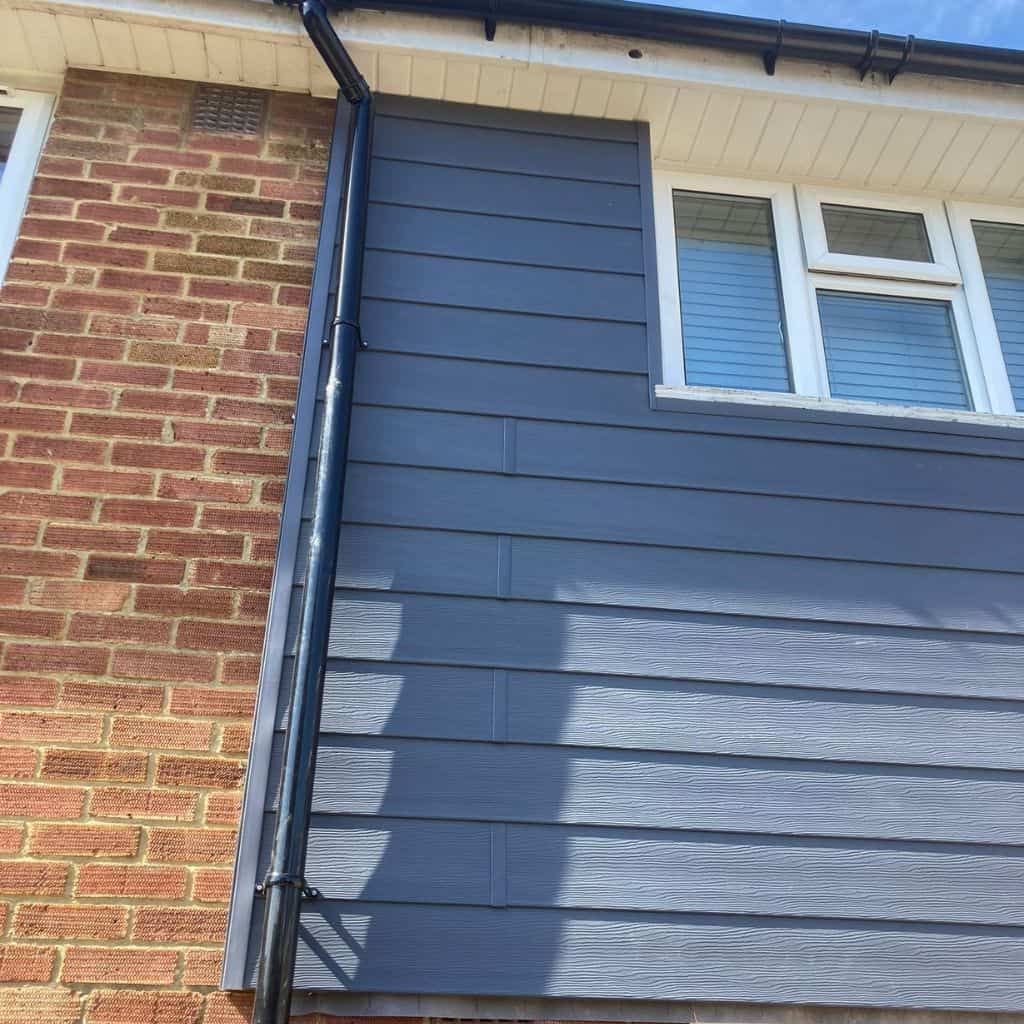 Grey fibre cement cladding fitted to a house wall beside red brickwork, with a black downpipe and uPVC window above.