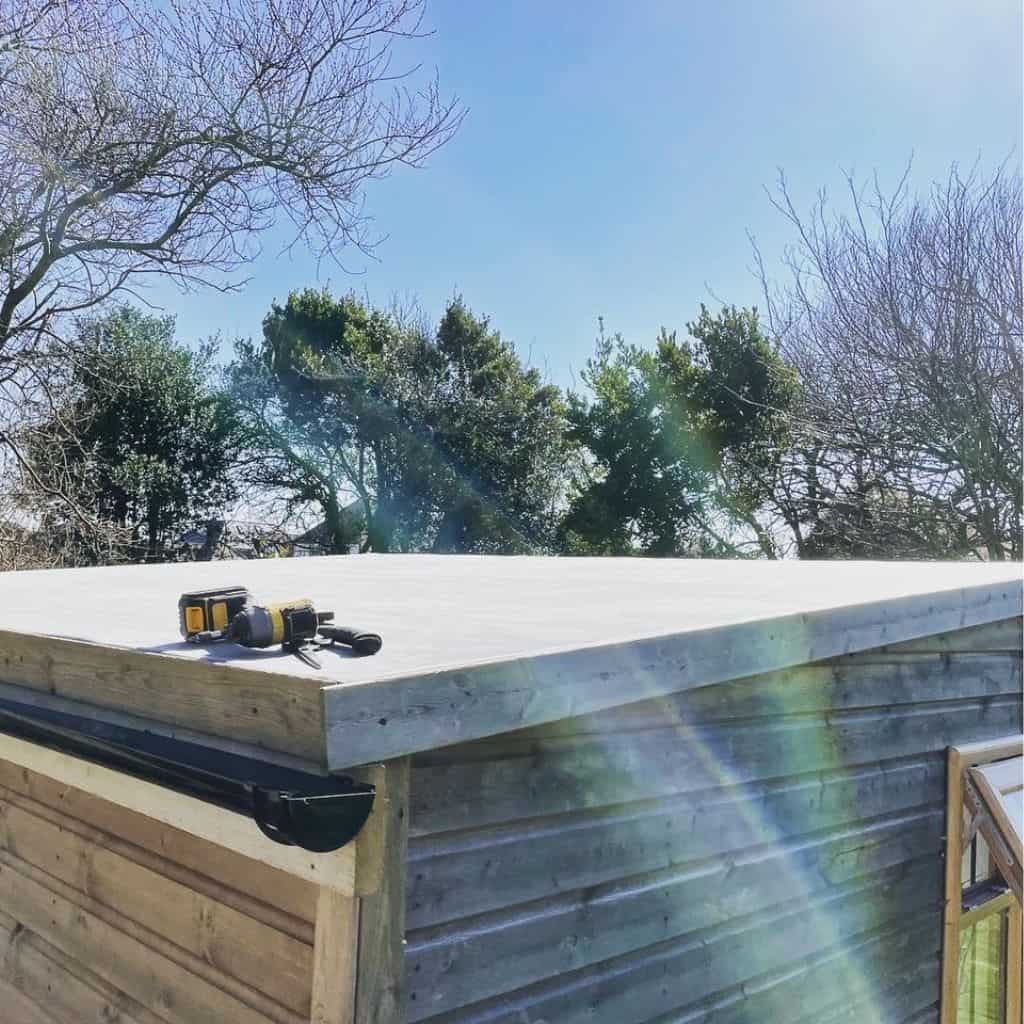 Newly fitted pale flat roof on a timber outbuilding with a black gutter and a cordless drill resting on the surface.
