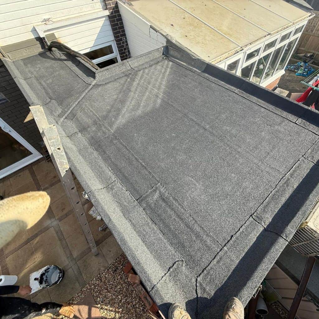 Newly laid grey felt flat roof on a domestic extension, edged with trims beside a conservatory.