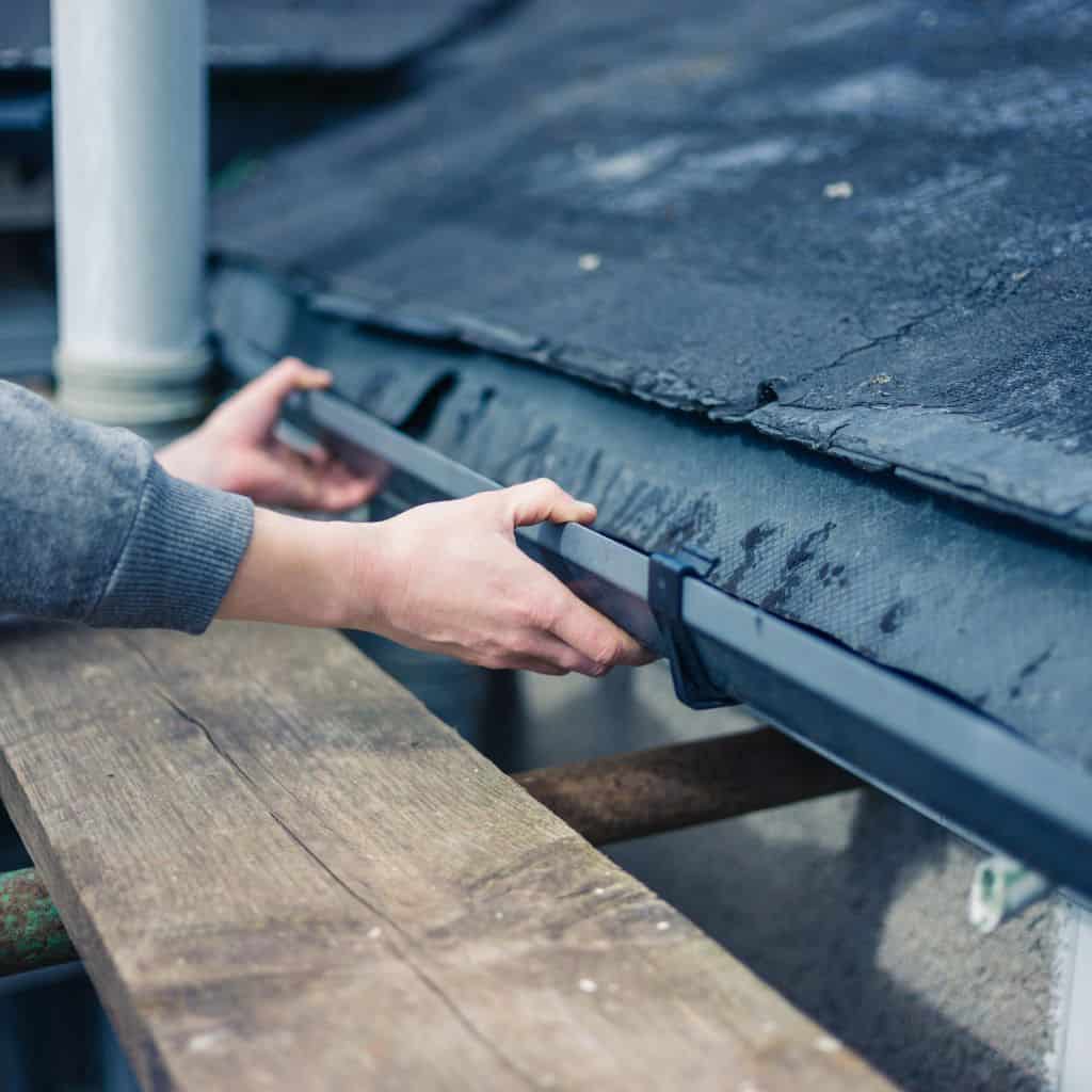 Roofer's hands inspecting the edge of a black felt flat roof and its guttering while standing on a scaffold board.