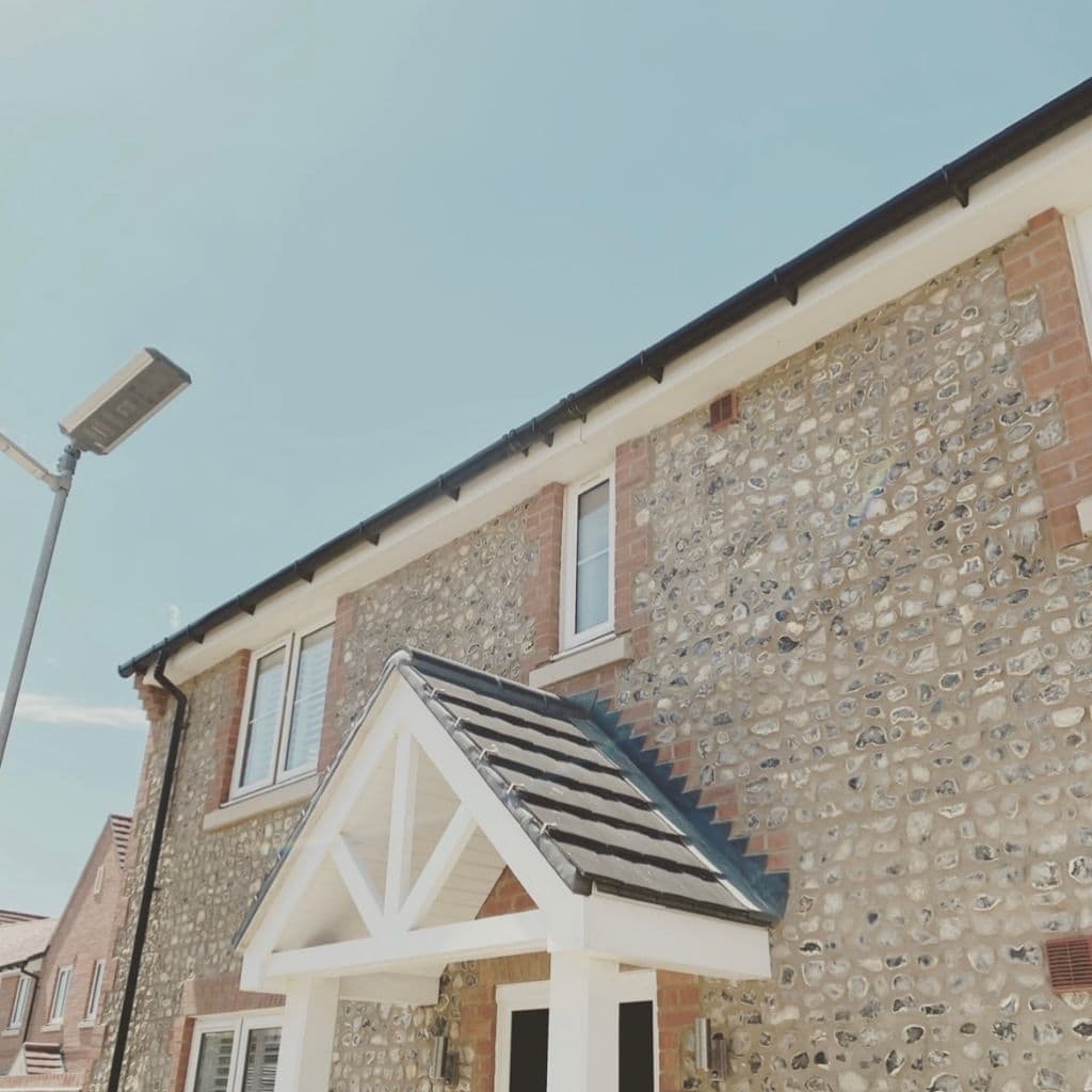 Black ogee guttering and round downpipe along the eaves of a flint and brick house under a clear blue sky.