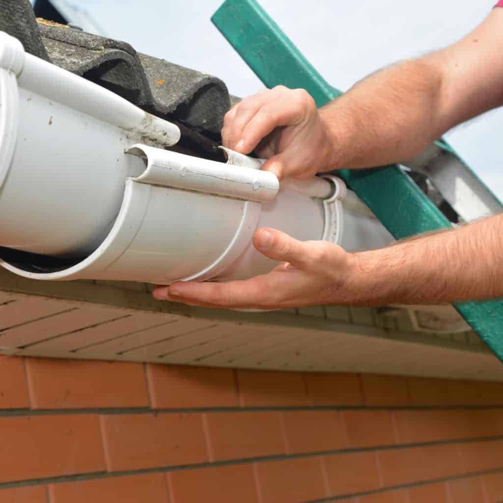 Close-up of hands fitting a white uPVC gutter section at the roof eaves, with a ladder resting against the wall.