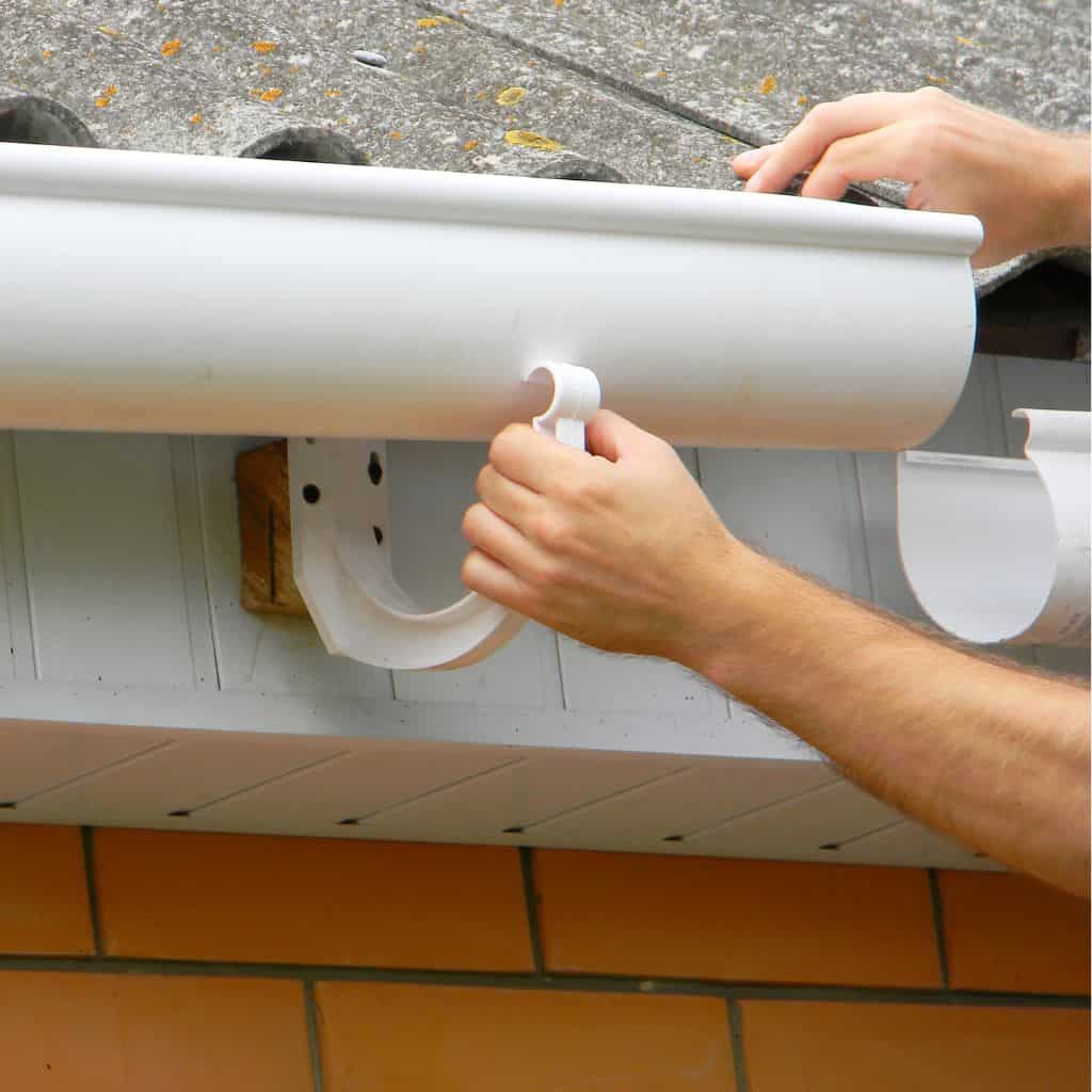Hands fitting a new white half-round PVC gutter onto fascia above an orange brick wall.