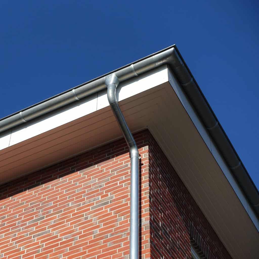Corner of a brick building with a galvanised steel gutter and downpipe beneath the eaves against a clear blue sky.