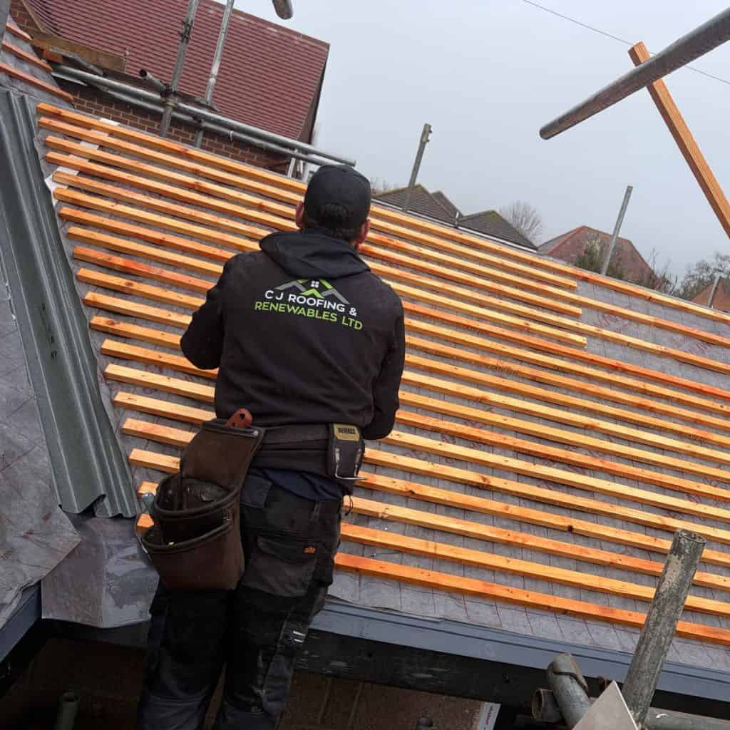 Roofer in a C J Roofing & Renewables hoodie fitting timber battens to a stripped pitched roof during reroofing.