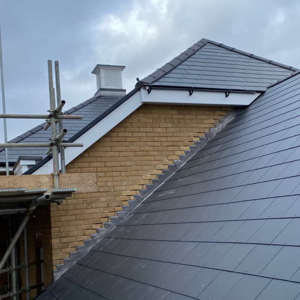 Pitched roof of dark grey slate tiles meeting a yellow brick gable wall, with scaffolding and a louvred vent above.