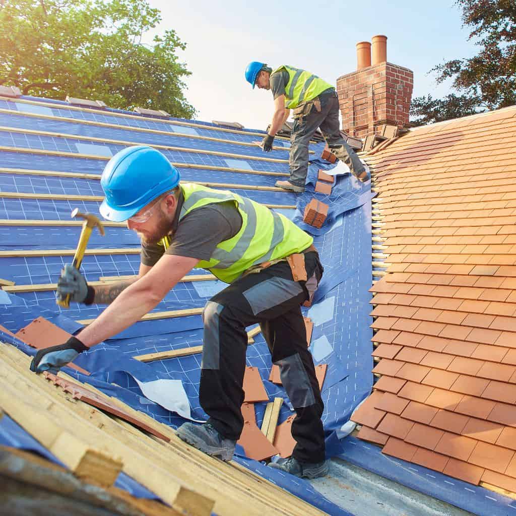 Two roofers in hi-vis and hard hats laying red clay tiles over blue underlay battens, working towards a brick chimney.