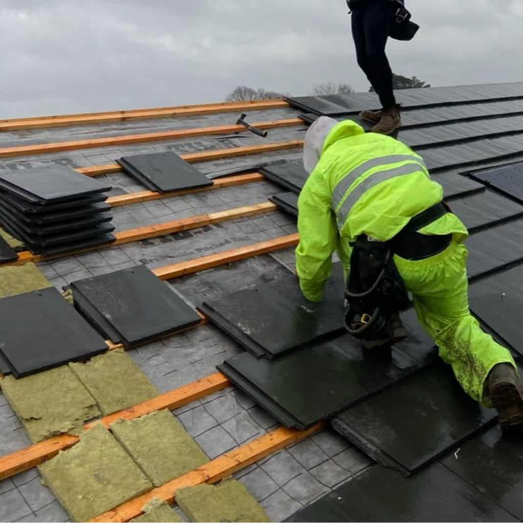 Roofer in hi-vis fixing dark slate-effect tiles to battens over breathable membrane on a pitched roof in wet weather.