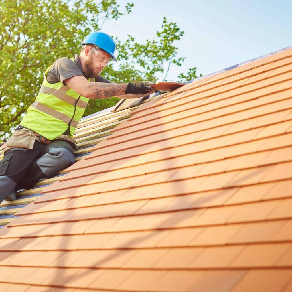 Roofer in a hi-vis vest and hard hat laying terracotta roof tiles on a sunlit pitched roof beside trees.
