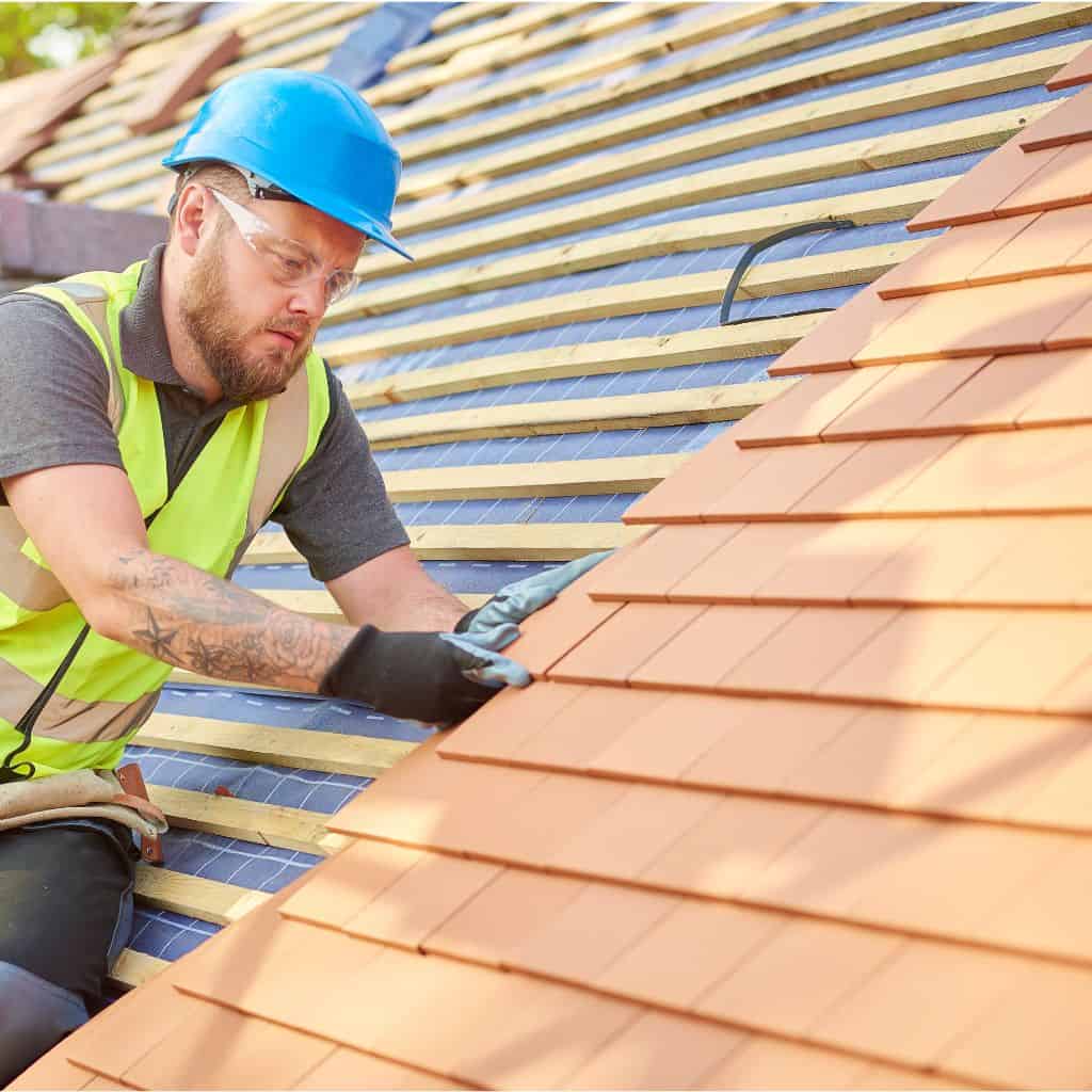 Roofer in a hi-vis vest and blue hard hat laying red clay tiles on battens up a pitched roof.