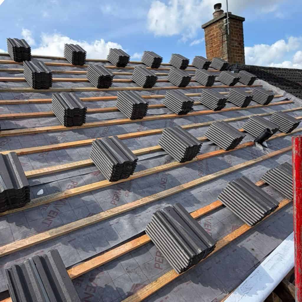 Bundles of grey concrete roof tiles stacked along timber battens over breathable membrane, beside a brick chimney.