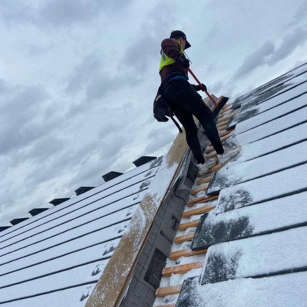 Roofer in a hi-vis vest sweeping light snow from the ridge of a tiled pitched roof under a grey sky.