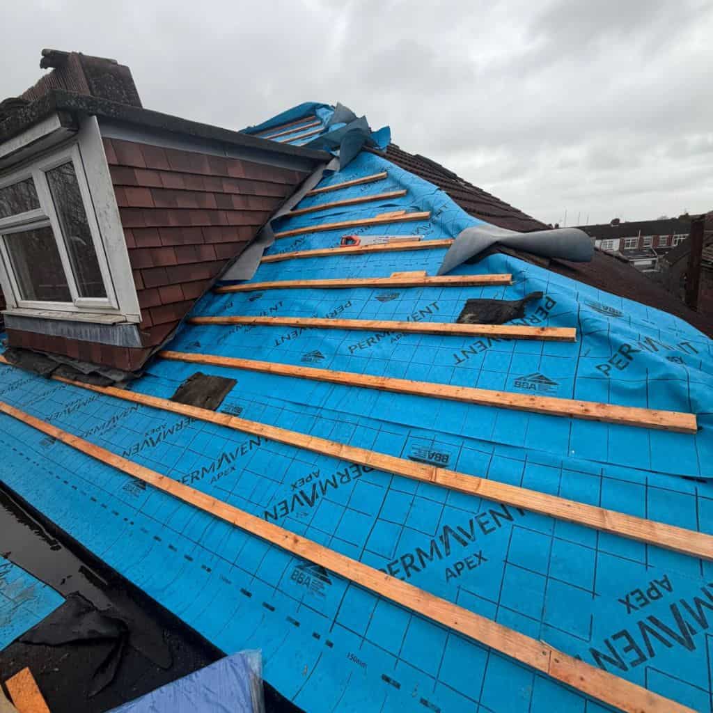 Pitched roof stripped to blue breathable underlayment with new timber battens being fixed, beside a tiled dormer window.