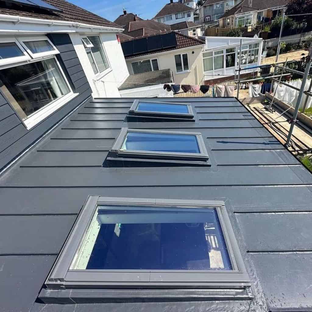 Three flat rooflight windows set into a dark grey flat roof membrane, with houses and gardens visible behind.